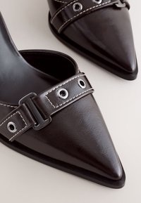 Chaussures en cuir noires à bout pointu avec des brides décoratives comportant des œillets métalliques et des surpiqûres sur un fond clair.