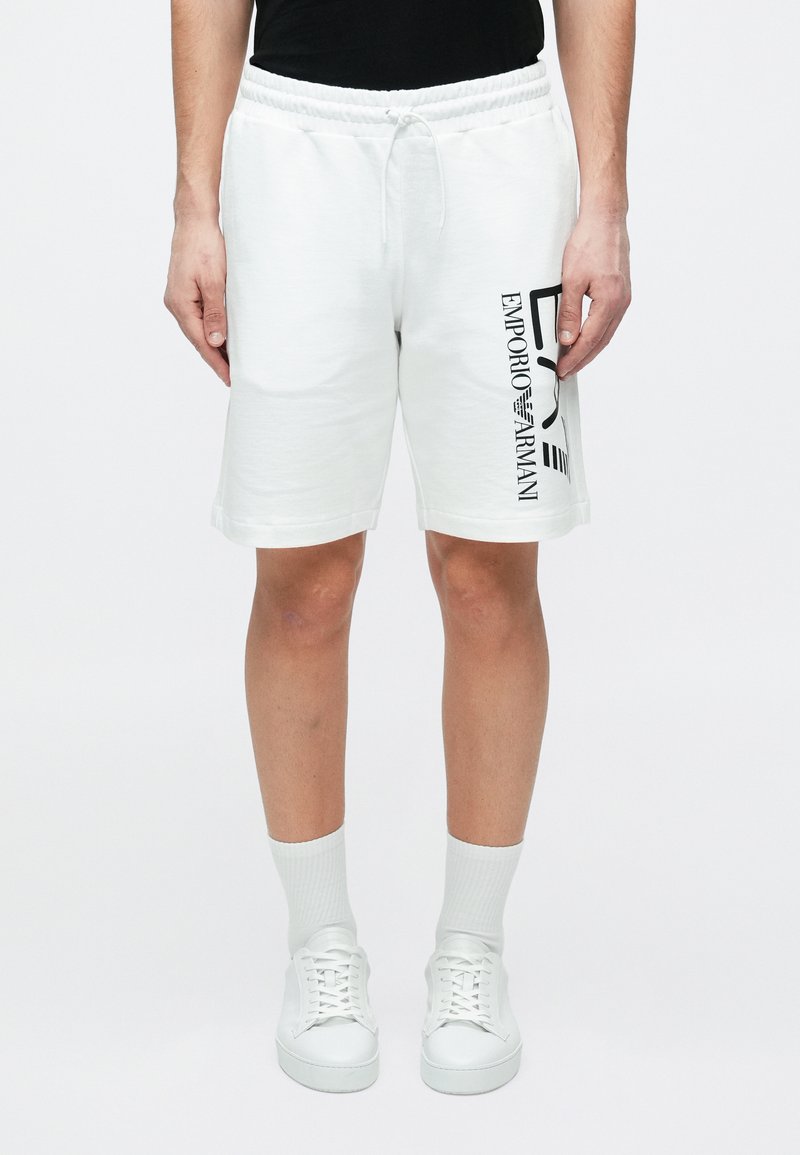 Άνδρας που φοράει λευκά σορτς Emporio Armani, λευκές κάλτσες και λευκά αθλητικά παπούτσια, στέκεται μπροστά σε ένα απλό λευκό φόντο.