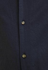 Camisa de un oscuro navy hecha de un tejido texturizado, con dos botones marrones a lo largo del placket delantero. Diseño simple y limpio.