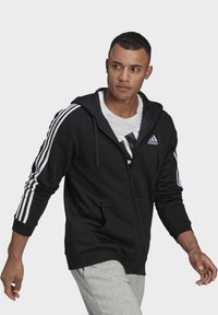 Sweatshirt preto com fecho éclair, feito de tecido macio, com destaques em branco em três riscas nas mangas e um bolso frontal. Inclui um capuz ajustável.
