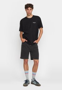 Zwarte T-shirt met korte mouwen en een klein wit logo, gecombineerd met donkergrijze denim shorts. Accessoires omvatten grijze sokken met rode en blauwe strepen en grijze sneakers.