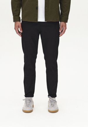 Black tapered pants with a smooth texture, paired with gray sneakers and an olive green button-up shirt over a white T-shirt.
