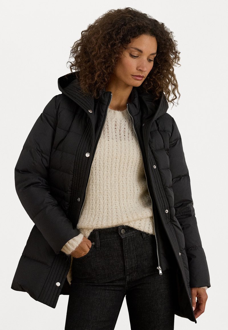 Μαύρο κοντό τζάκετ με επένδυση και κουκούλα, που διαθέτει φερμουάρ και κούμπωμα με κουμπιά, φορεμένο πάνω από ένα ανοιχτό πλεκτό πουλόβερ και σκούρα τζιν.