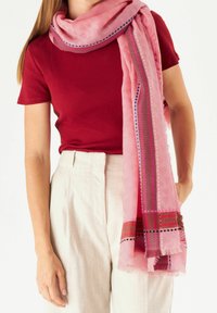 Écharpe rose clair avec un motif texturé et des bordures colorées présentant des accents bleus et rose foncé. Portée lâche autour du cou.