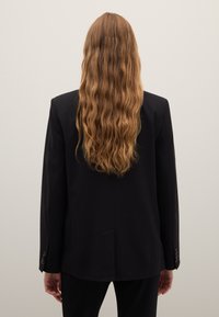 Blazer in lana nera con una vestibilità su misura, caratterizzato da maniche lunghe, spacco singolo sul retro e dettagli con bottoni sui polsini. Capelli lunghi e ondulati visibili.