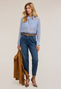 Camicia azzurra chiara trasparente con bottoni e due tasche sul petto, jeans in denim a vita alta, cintura marrone e giacca in pelle beige appoggiata sul braccio.
