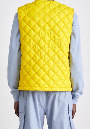 Personne portant un gilet matelassé jaune vif sur une chemise à manches longues bleu clair et un pantalon bleu clair avec des poches cargo latérales, vue de dos.