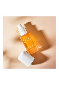 Bernsteinfarbene Glasflasche mit einem weißen Pumpverschluss, beschriftet mit "rms SuperSerum Hydrating Mist", neben einem weißen zylindrischen Deckel auf beiger Oberfläche platziert.