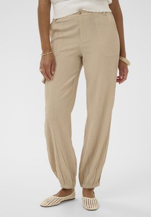 Beige textuurbroek met elastische boorden, gedragen met witte gevlochten platte schoenen en een wit mouwloos topje, aangevuld met armbanden en ringen.