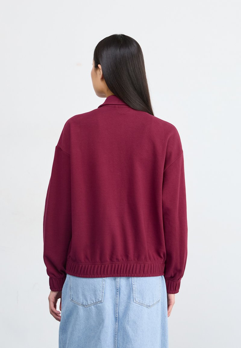 Burgunderfarbene Sweatshirt mit geripptem Saum und Bündchen, mit einem lockeren Schnitt und einem umklappbaren Kragen. Getragen über einem hellblauen Jeansrock.