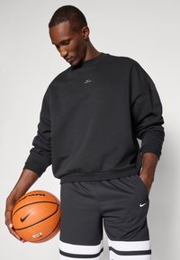 Sort crewneck sweatshirt med ribbede manchetter, parret med mørkegrå shorts med hvide striber. Modellen holder en orange basketball.
