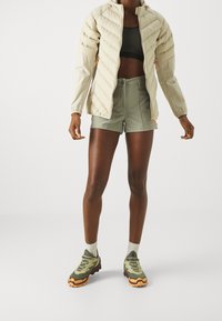 Ljusbeige pufferjacka med horisontell quiltad mönster, matchad med olivgröna shorts och kraftiga gröna-orange vandringskor.