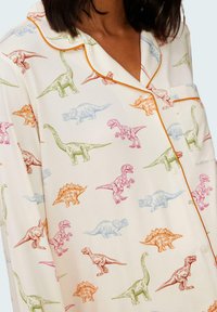 Vitt tygpysset med färgglada dinosauriemönster i rosa, grönt, blått och orange, med kontrasterande orange kantning och fickdetalj.