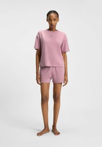 Una giovane donna in piedi, a piedi nudi, che indossa un abbigliamento coordinato composto da una maglietta rosa ampia e dei pantaloni shorts, su uno sfondo neutro.