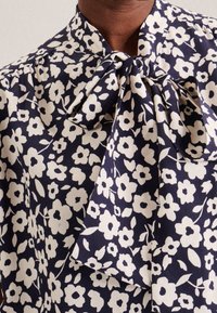 Mörkblå blus med vitt blommönster, med en detaljerad knytning vid halsen. Tyget verkar lätt och har en mjuk struktur med en subtil lyster.