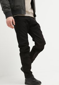 Svarta byxor med dragkedjeförsedda framfickor, slimmad passform och elastisk midja. Kombineras med en grå bomberjacka och svarta skor.