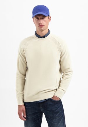 Beige gebreide trui met ribbelaccenten op de schouders, gecombineerd met een blauwe pet en donkere spijkerbroek. De trui heeft een ronde hals en lange mouwen.