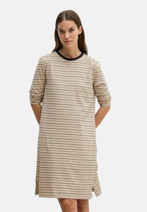 Vrouw draagt een beige en zwart gestreepte jurk met lange mouwen en een zwarte kraag, staat met haar handen achter haar rug tegen een effen achtergrond.