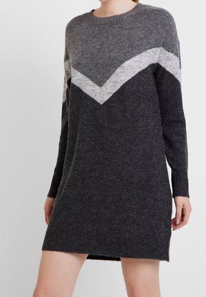 Frau trägt ein dunkelgraues Strickkleid mit einem hellgrauen Chevron-Muster über Brust und Armen vor einem einfarbigen Hintergrund.