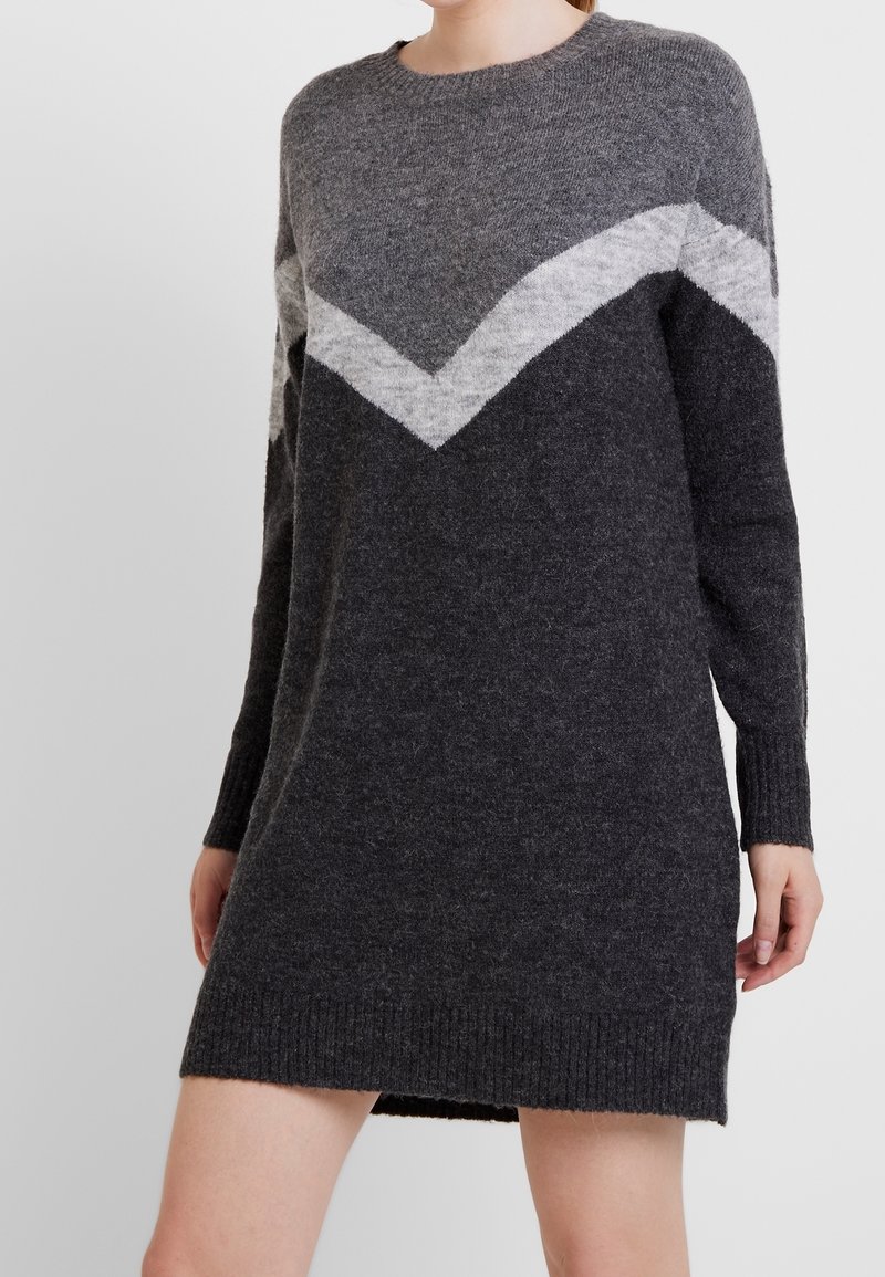 Femme portant une robe-pull tricotée gris foncé avec un motif chevron gris clair sur la poitrine et les bras sur un fond uni.