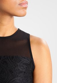 Robe noire sans manches avec une partie supérieure en dentelle transparente et des détails en dentelle à motifs au niveau de la taille. Texture lisse et design ajusté.