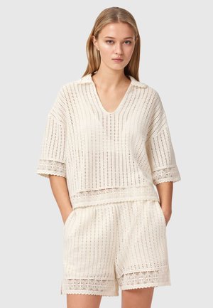 Ung kvinde iført cremefarvet strikket kortærmet top med krave og matchende strikkede shorts med blonderkant, stående med hænderne i lommerne.