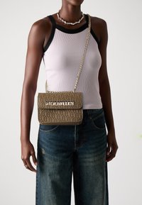 Beige skulderveske med preget logo-mønster, hardware i gulltone og kjede-rem, holdt av en person som har på seg en ribbet tanktopp og jeans.