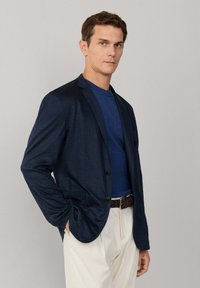 Muž v námorníckom saku, modrej košeli, bielych nohaviciach a hnedom opasku stojaci s jednou rukou v vrecku na jednoduchom pozadí.