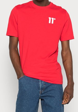 Man draagt een felrode T-shirt met korte mouwen met een wit "11°"-logo op de borst en een blauwe spijkerbroek.
