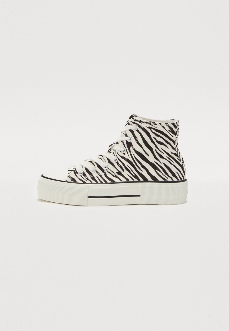 Høye sneakers i hvit lerret med et svart zebra-mønster, med hvite snørebånd og en tykk gummisåle med et svart stripeakcent.