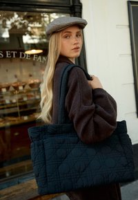 Borsa a tracolla nera trapuntata con motivi a rombi, texture in tessuto e manici robusti. Un modello che indossa un maglione marrone scuro si trova davanti a una panetteria.