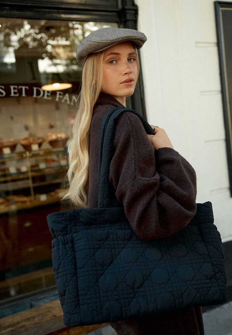 Borsa a tracolla nera trapuntata con motivi a rombi, texture in tessuto e manici robusti. Un modello che indossa un maglione marrone scuro si trova davanti a una panetteria.