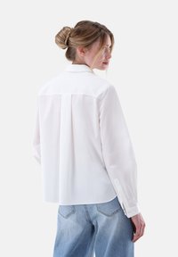 Camicia bianca a maniche lunghe con vestibilità comoda, caratterizzata da una piega sul retro e polsini con bottoni, indossata con jeans di denim azzurro chiaro.