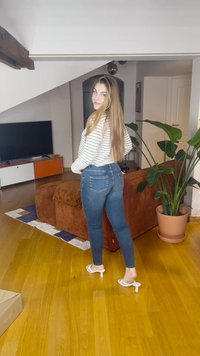 Kék farmer sztreccs szűk nadrág fehér hosszú ujjú csíkos felsővel és fehér magas sarkú szandállal párosítva, otthonos beltéri térben egy növénnyel.