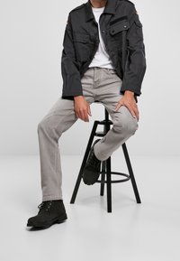 Chaqueta negra de lona con dos bolsillos en el pecho, jeans de mezclilla gris y botas de ante negras hasta el tobillo, sentado en un taburete negro frente a un fondo liso.