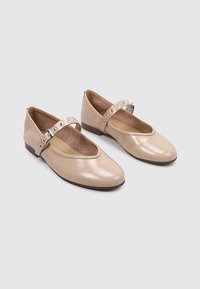 Zapatos de charol beige con puntera redonda, suela plana y una delgada correa en el tobillo con tachuelas circulares plateadas.