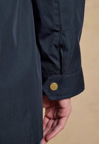 Nahaufnahme eines Ärmelbündchens einer dunkelblauen Jacke mit einem goldfarbenen Druckknopf, der einen Teil einer entspannten Hand vor beigem Hintergrund zeigt.