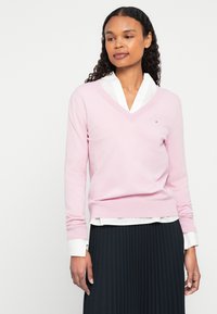 Maglione rosa a scollo a V in morbido tessuto a maglia, con polsini e orlo a coste. Indossato sopra una camicia bianca con colletto, abbinato a una gonna nera plissettata.