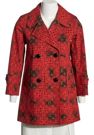 Abrigo rojo de doble fila con botones negros, con un patrón de círculos rojos y logotipos monograma marrones, exhibido en un maniquí blanco.