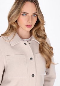 Beige wool-blend jacket featuring a collar, two front pockets, and dark buttons. Smooth texture with a relaxed fit, suitable for layering.