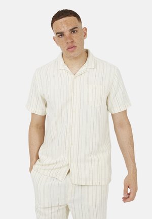 Młody mężczyzna o krótkich włosach, ubrany w kremową koszulę na krótki rękaw na guziki oraz pasujące spodnie w paski, stojący z jedną ręką w kieszeni.