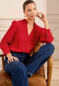 Blusa rossa con cuciture decorative e scollatura a V. Maniche lunghe con polsini arricciati, abbinata a jeans blu scuro. Seduta su una sedia di legno.