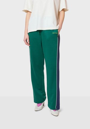 Pantalons de survêtement verts avec des panneaux latéraux navy et un passepoil blanc, dotés d'une taille élastique. Associés à une chemise ample couleur crème et des chaussures blanches.