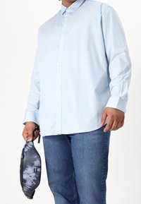 Camicia azzurro chiaro a maniche lunghe con bottoni, in cotone, abbinata a jeans di denim scuro. Tienendo una borsa a tracolla con motivo blu e grigio.