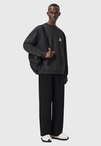 Felpa in cotone nera con logo frontale, abbinata a pantaloni neri. Il modello porta uno zaino nero testurizzato. Sneakers bianche completano l'aspetto.