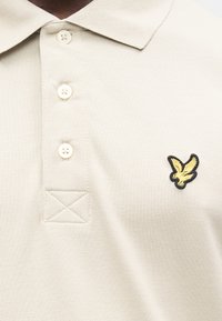 Polo color beige realizzata in tessuto strutturato con un colletto a tre bottoni. Presenta un logo di aquila ricamato in nero e oro sul petto.