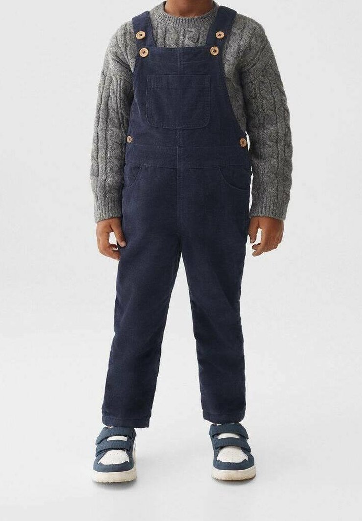 Enfant portant une salopette bleu marine avec des boutons en bois, un pull en maille torsadée gris et des baskets bleues et blanches à velcro, debout sur un fond uni.