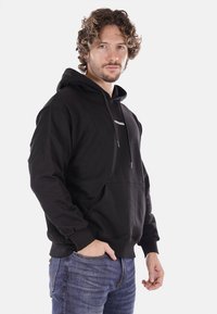 Felpa nera in cotone con tasca a marsupio, con cordini, e un logo bianco sul petto, abbinata a jeans blu.