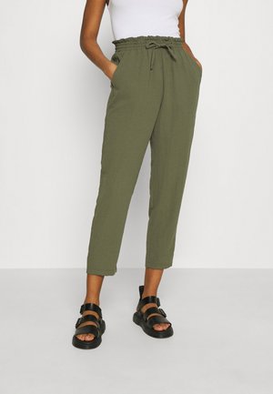 Femme portant un pantalon court taille haute couleur vert olive avec un cordon de serrage et des sandales noires à boucle, debout les mains dans les poches.