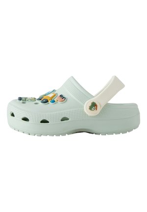 Svijetlozelena udobna natikača s rupicama za ventilaciju, podesivim remenom na peti i šarenim ukrasnim privjescima na vrhu.